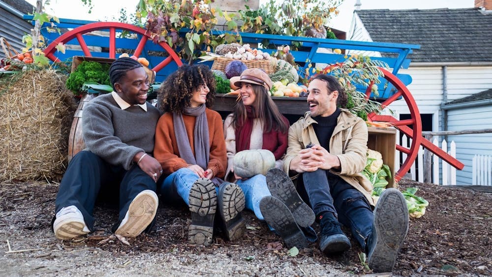 Group of four friends sitting on ground in front of a wagon filled with autumn produce and hay bales