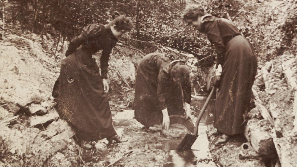 Three people in feminine attire panning for gold in a stream