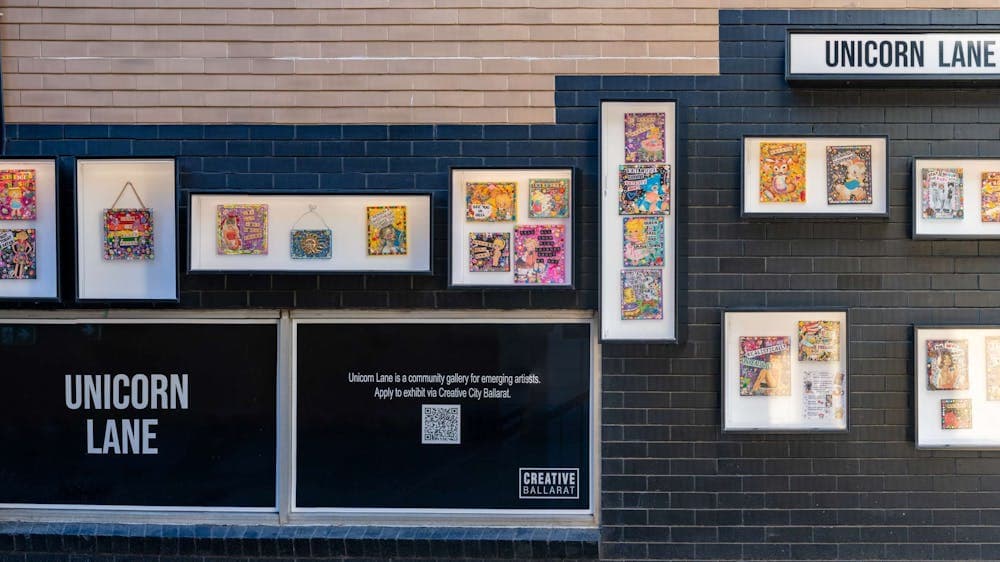 Full display of outdoor exhibition boxes filled with mixed media works on a black brick wall.