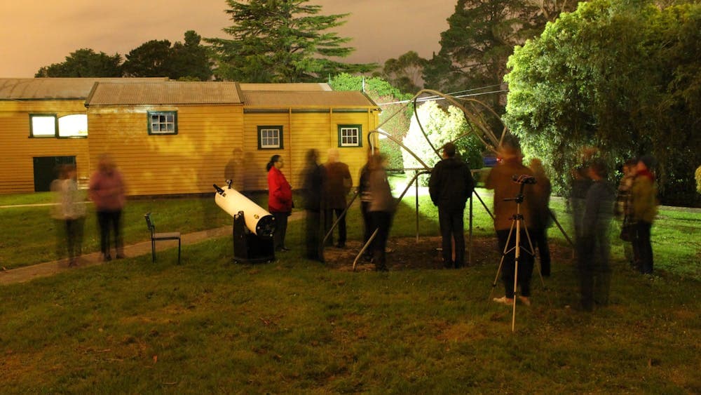 Small group stands around, telescope, binoculars and pipeghenge on grass in front of yellow building