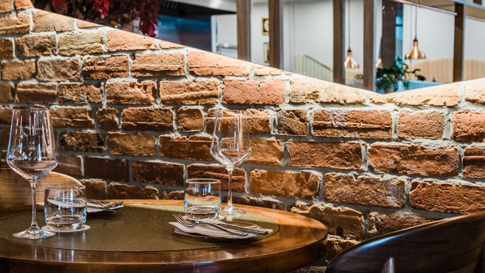 A dining table against a brick wall, set with cutlery and wine glasses