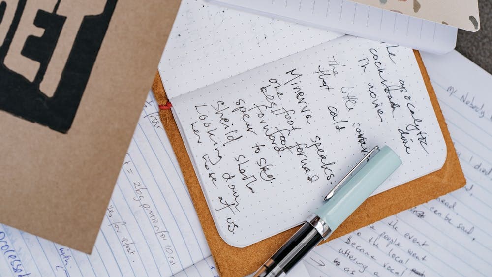 Several notebooks, one in the centre laying open to reveal a handwritten poem, and a light blue pen