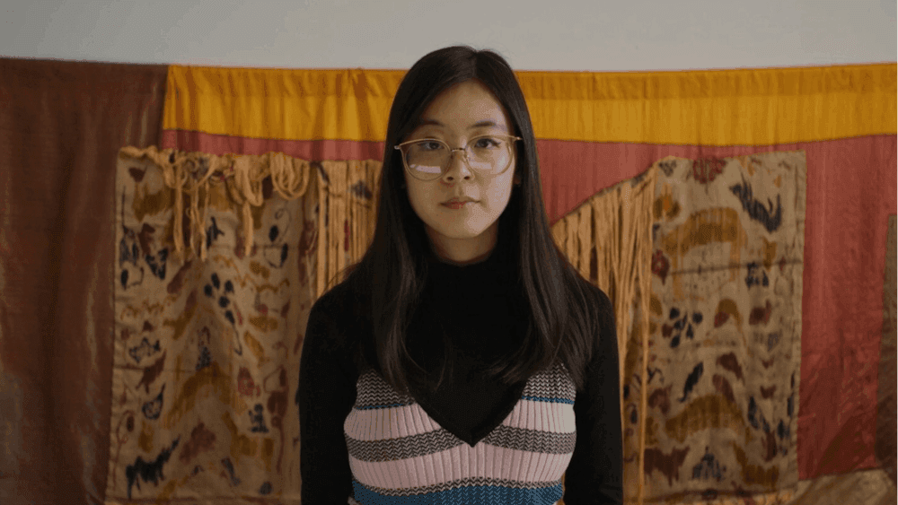 Woman in black attire with printed top standing in front of a wall weaving