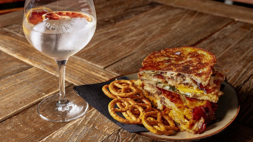 A tall glass of ice cold gin and tonic nect to a toasted sandwhich, served with pretzels