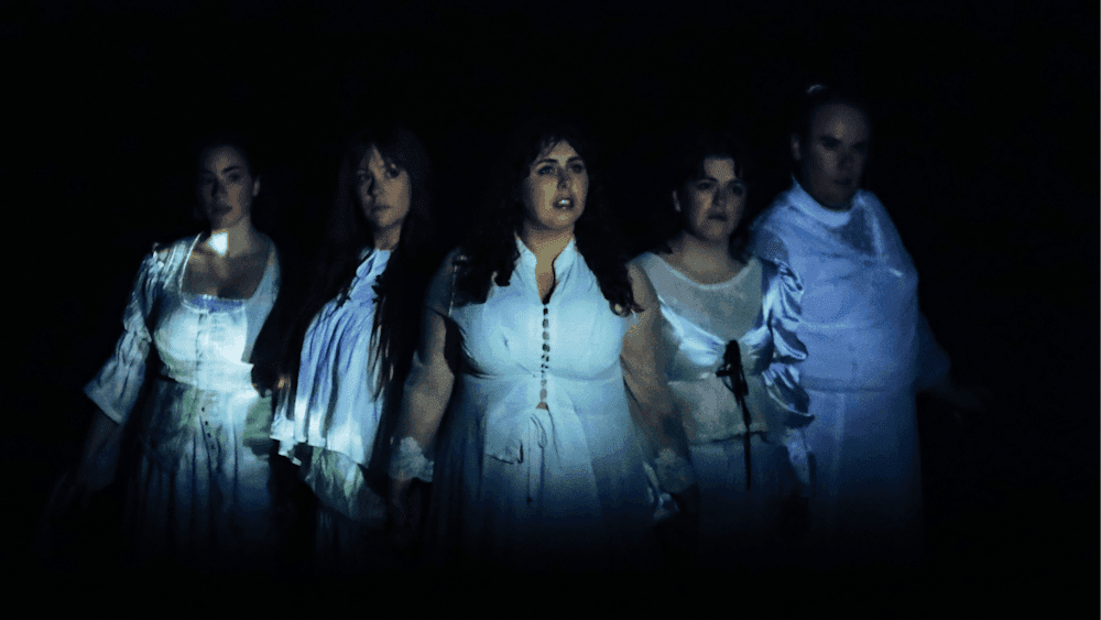 Five women dressed in white, holding hands and looking out to the distance.