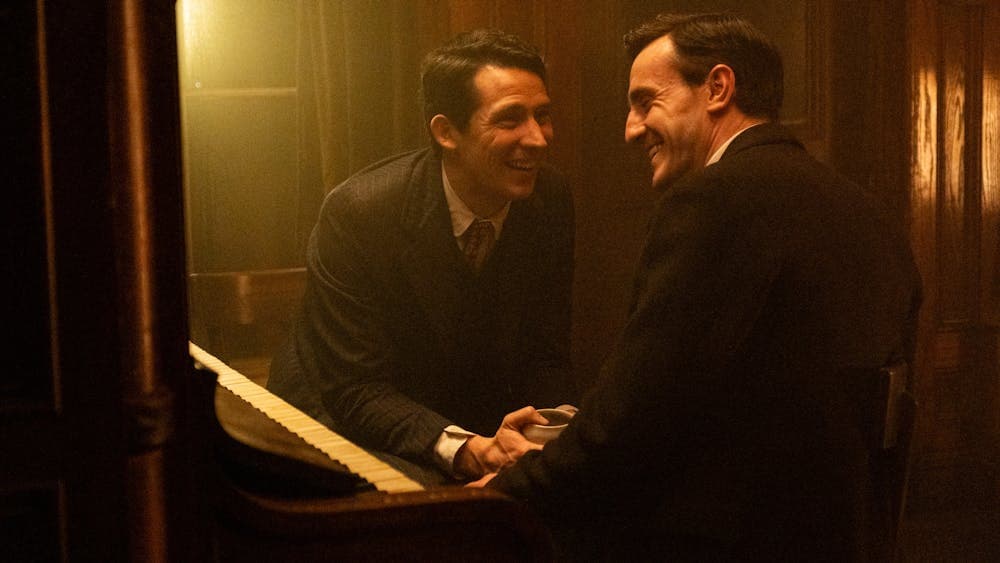 Two men lean on a piano while laughing