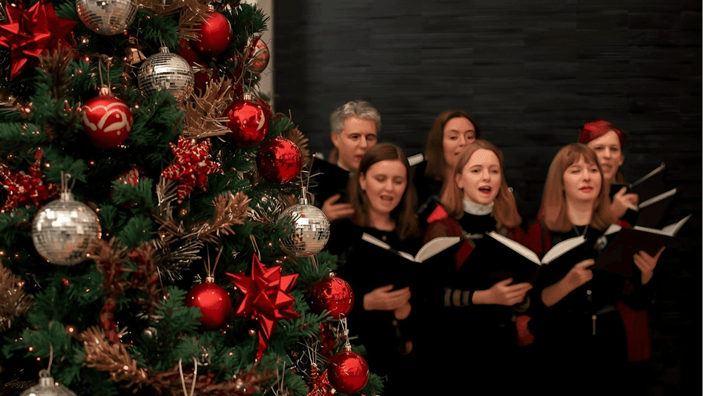 A group of carollers gathered around a Christmas tree singing. The  tree has red and mirror baubles