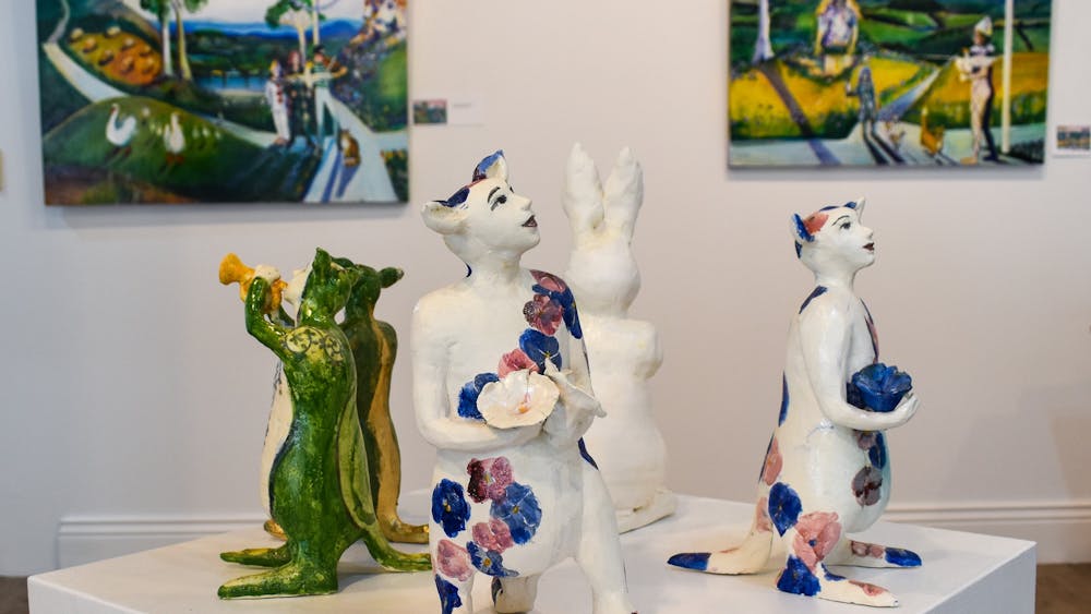 Landscape paintings in the background with whimsical ceramic characters on a plinth in the front.