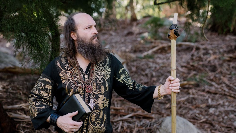 A man with a beard in intricate clothing, holding a book and staff