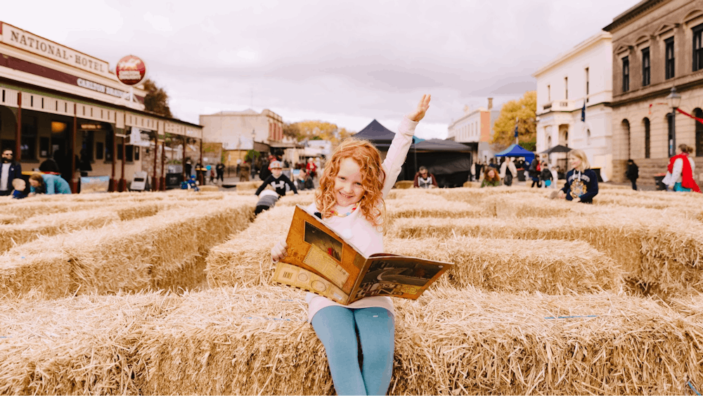 Girl sitting on hay bale reading a book with one arm in the air