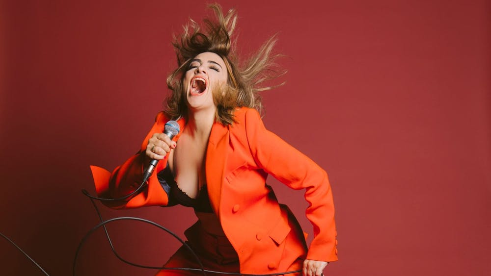 Woman in an orange suit howling into a microphone against a red background