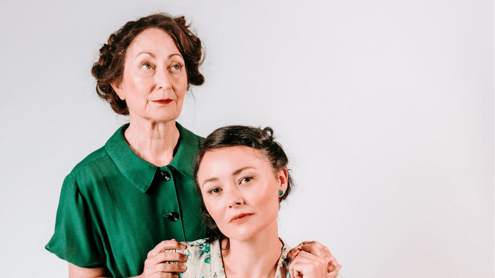 An older woman pensively rests against a younger woman, both dressed in 1953 attire