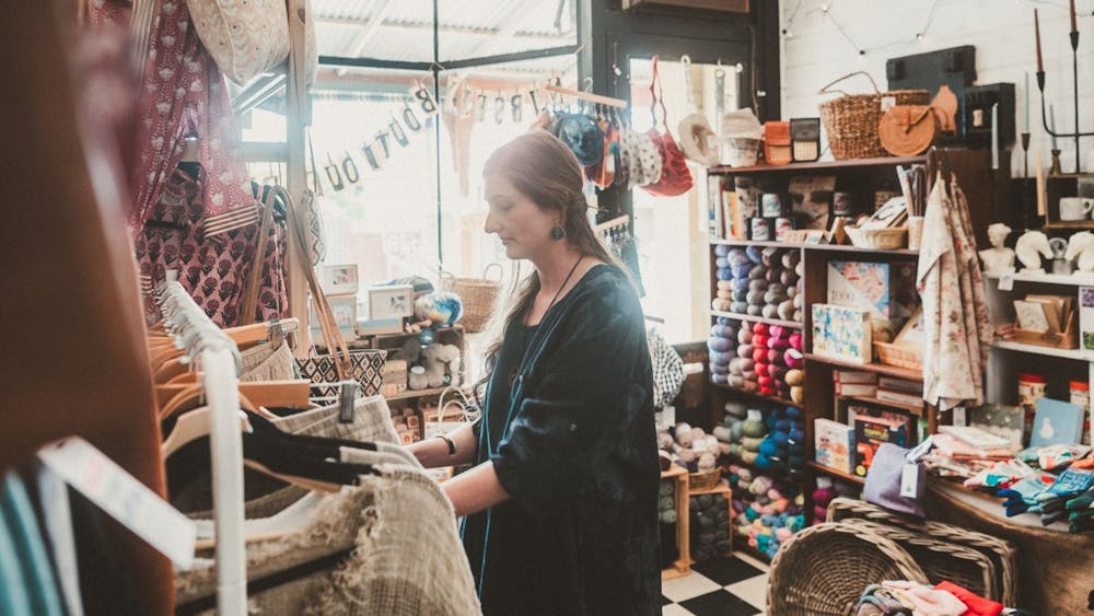 A woman in a blue dress looking at a rack of clothing, behind her is balls of wool and crafts