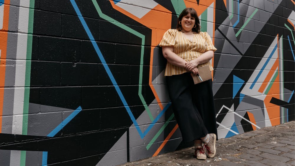 A fat woman with brown hair, black pants and a yellow top stands in front of a geometric shape mural
