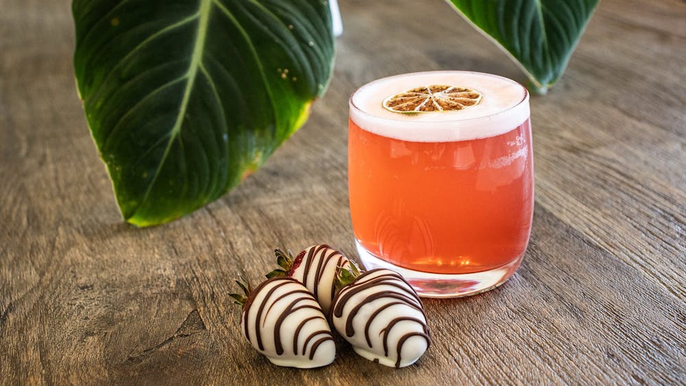 A pink cocktail in a short glass. on the left sits three chocolate covered strawberries