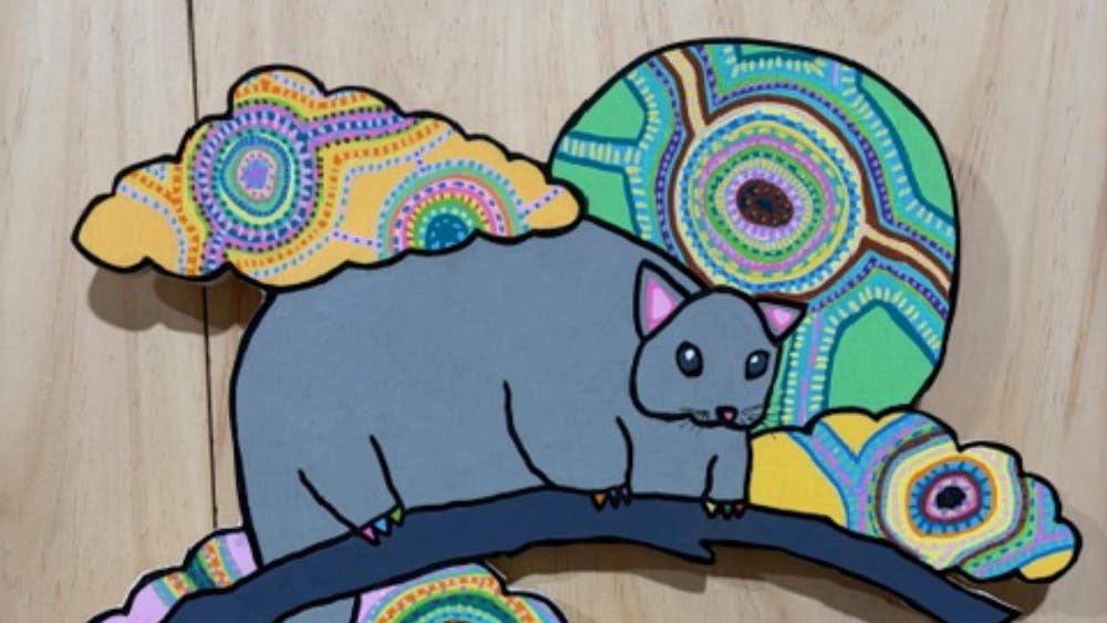 A wood cut brightly painted grey possum surrounded by brightly coloured flora.