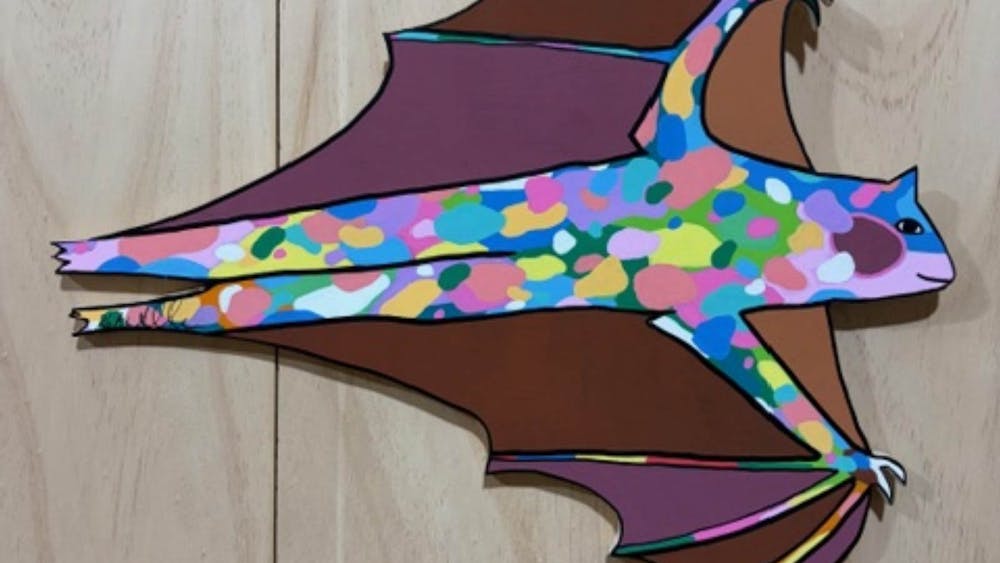 A flying bat made from cut wood and brightly painted on a wood background.