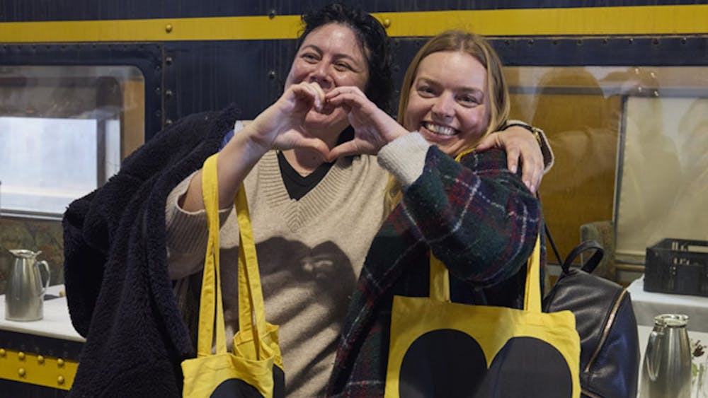 Two women making a heart shape by joining their hands. They are holding yellow and black tote bags.
