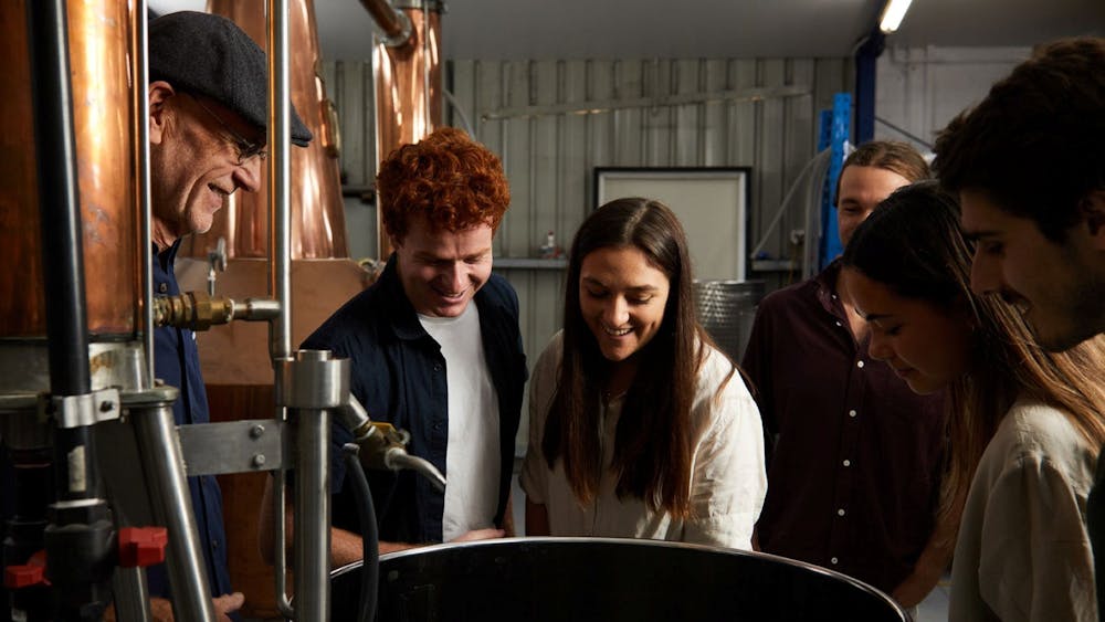 A small group of people peer into a large vat attached to a still.