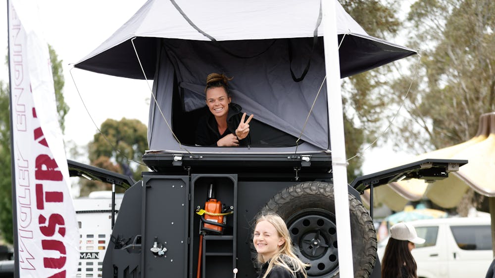 One young women lying in rooftop tent giving peace sign and  a second down below with a lollipop