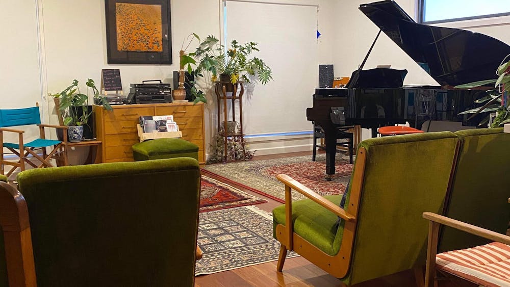 A black grand piano sits in front of lounge chairs; colourful rugs are on wooden floorboards.