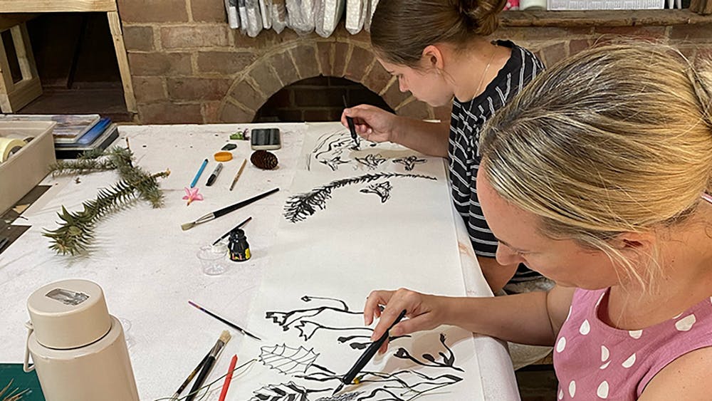 Two people drawing botanical forms in ink at a workshop table, historic brick fireplace behind.