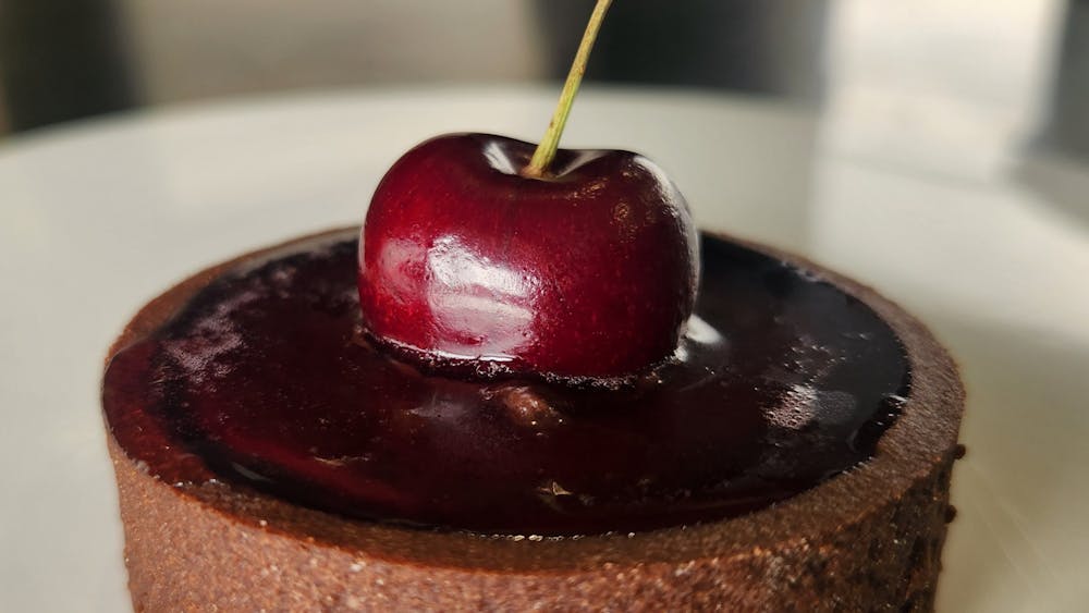 A chocolate tart sits on a white plate, it has a cherry jelly filling with a fresh cherry  on top
