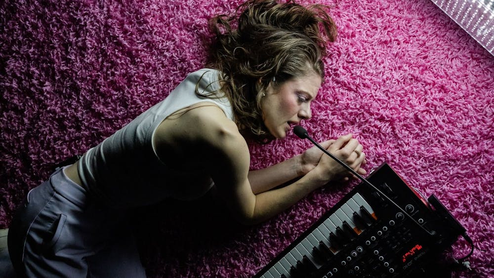 Karin McCracken looking forlorn, curled up on a pink carpet singing into a synth