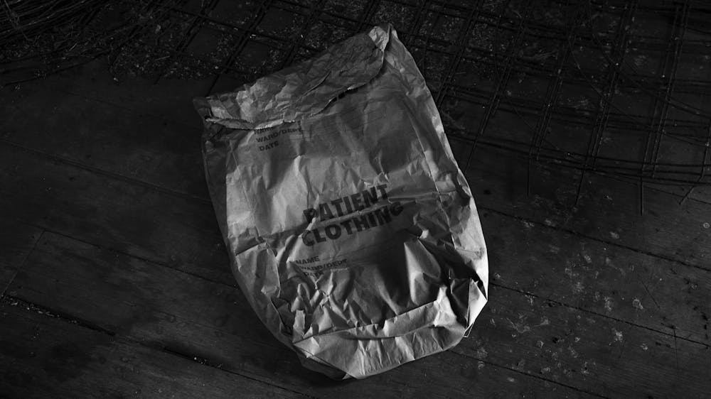 Black and white photograph of a crumpled hospital patient clothing bag on a wooden floor.
