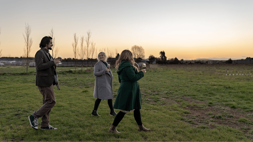 Three people walking holding wine glasses in a winery at sunset