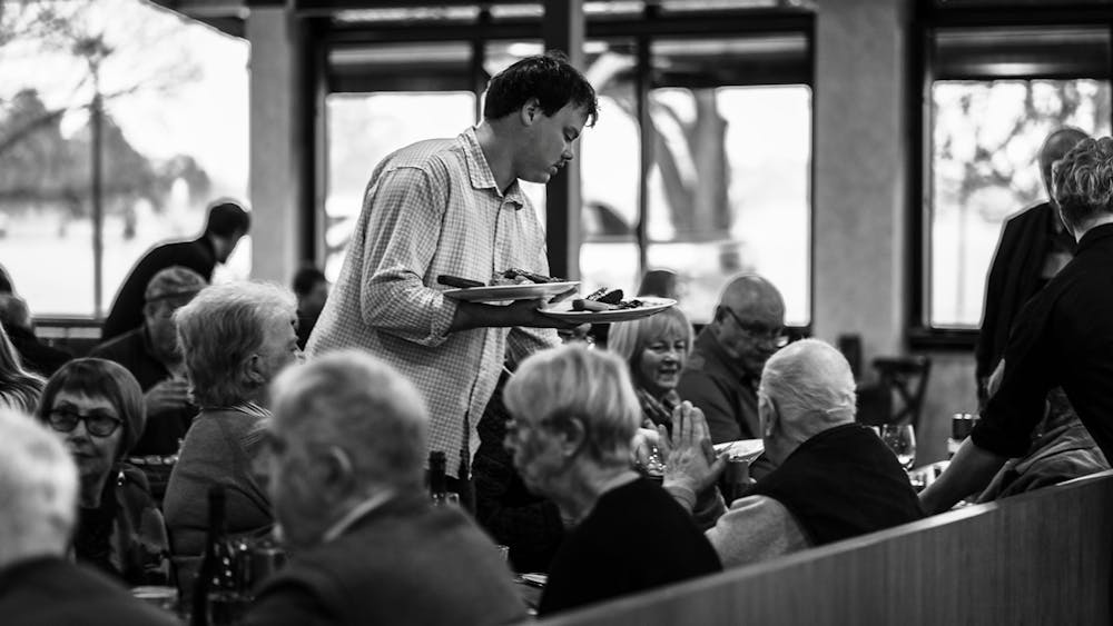 Staff member serving meals in a busy dining room filled with guests enjoying dinner together.