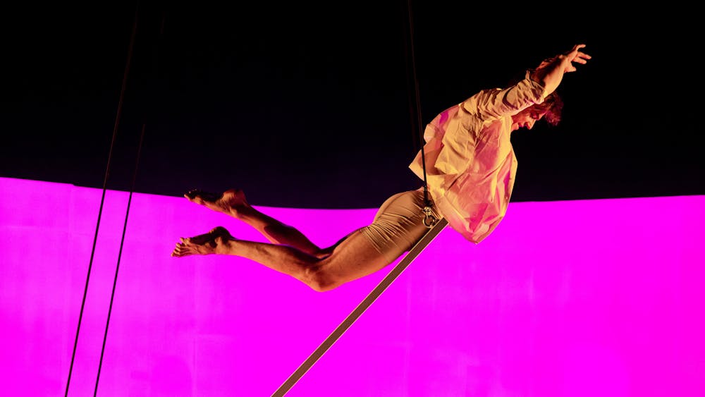A performer wearing an open shirt balances on an inclined board in a joyous moment of flight