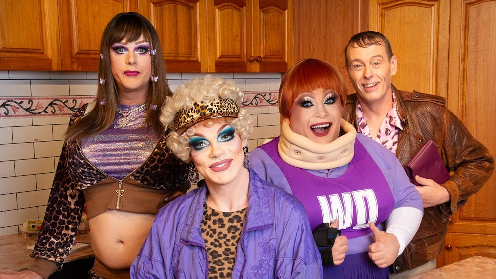 Four performers in colourful Y2K-inspired costumes and theatrical makeup pose together in a kitchen,
