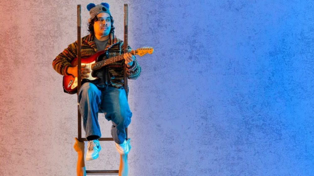 A musician sits at the top of a ladder playing a guitar. The ladder is being held upright in the air