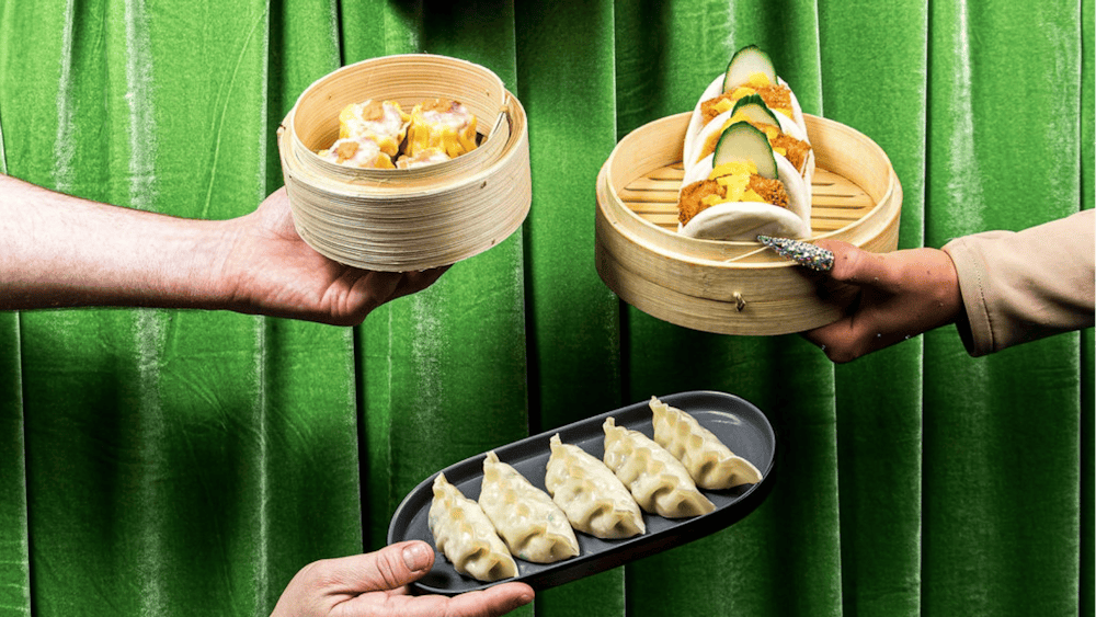 Hands hold bamboo steamers of bao and dumplings against a green curtain.