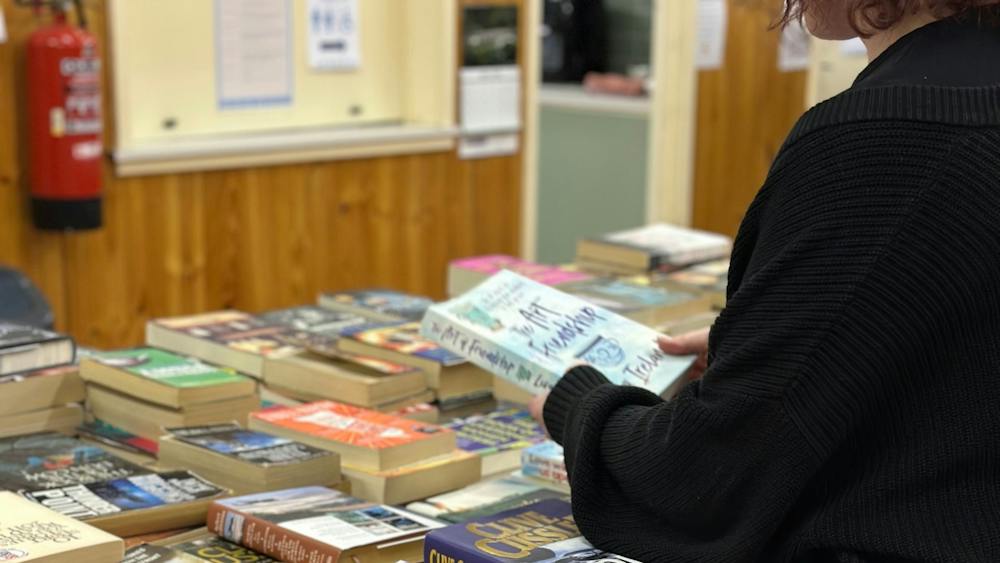 A person wearing a black cardigan holds a book while looking at a table stacked with books