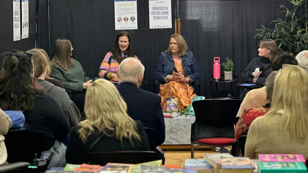 Background features four women talking about romance books, the audience blurred in the foreground.