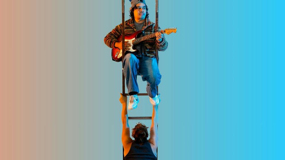 A guy in a beanie playing guitar on a ladder that is being held up by a muscular acrobatic woman
