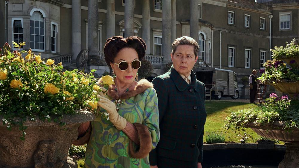 Two women stand in manor house garden next to yellow roses