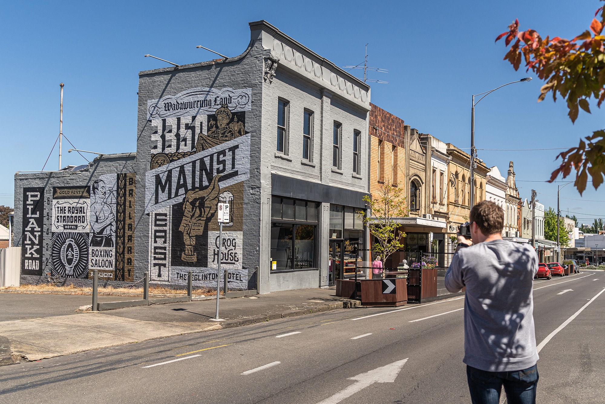 A man with his back to the viewer takes a photograph of the Main Road heritage streetscape and focuses on a mural on a building side wall by illustrator Travis Price.