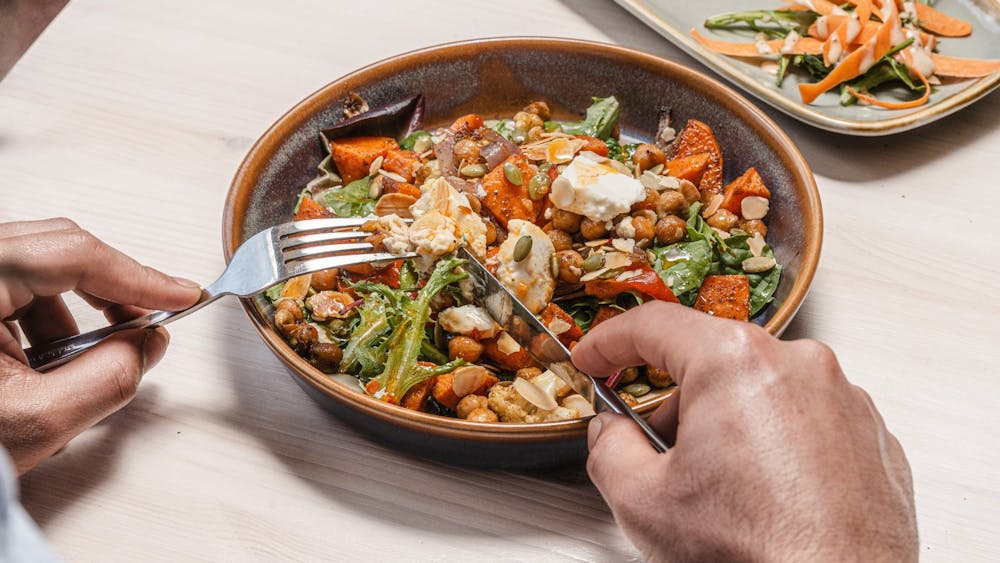 View of hands holding knife and fork, over a bowl of roast vegetable salad