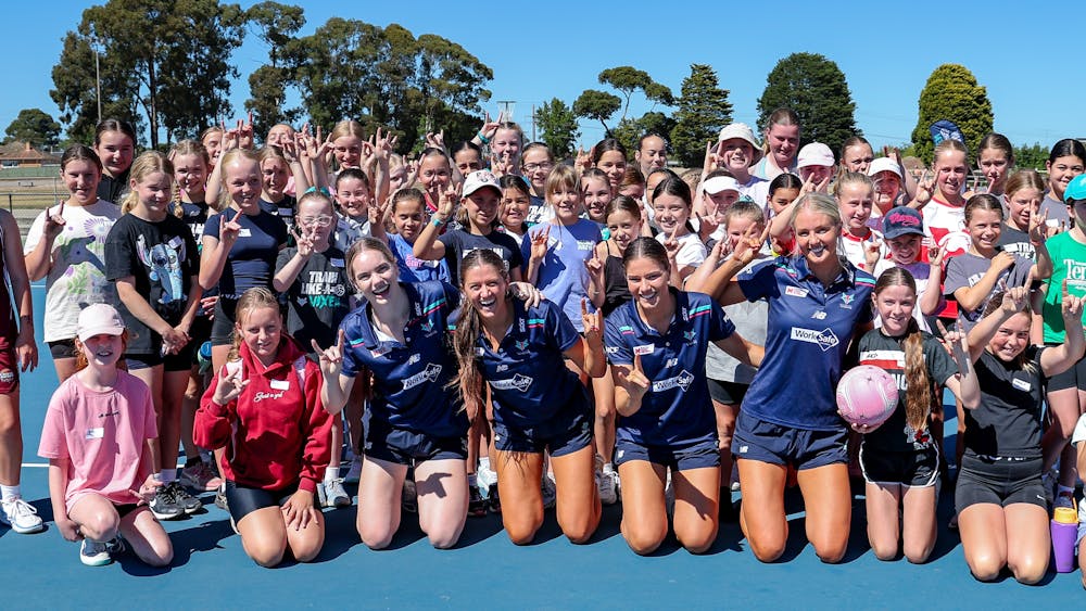 Four elite netballers lined up on a netball court with a group of excited young fans