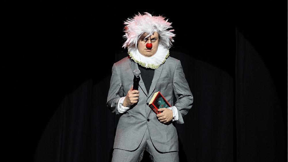 Artist in grey suit with white afro wig , red nose and white frilly color