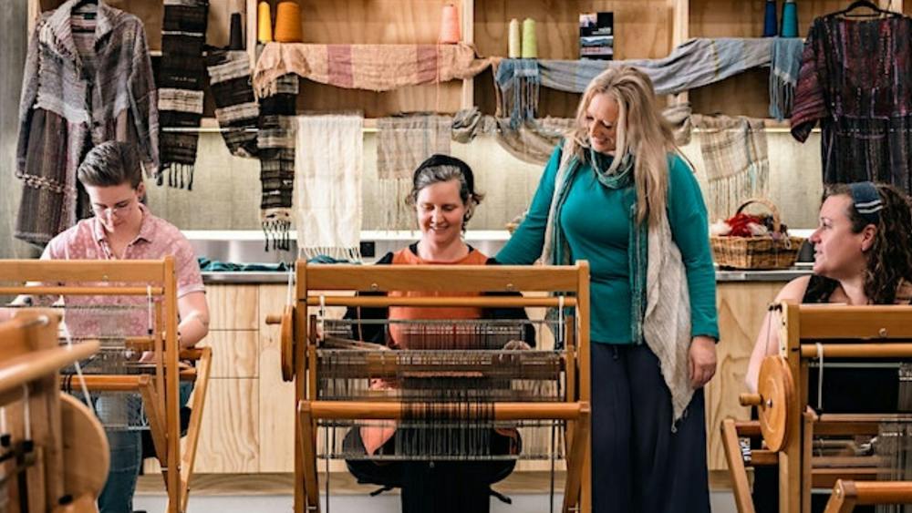 Three women seated at looms with instructor standing between them inside workshop