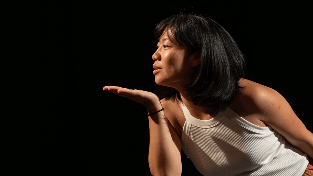 A dancer in a white sleeveless top and loose brown trousers leans forward under a spotlight.